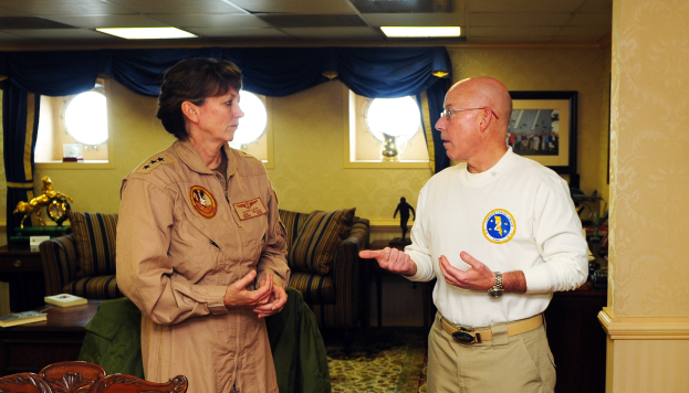 Ein Mann und eine Frau unterhalten sich in einem Wohnzimmer mit Möbeln, Büchern und einem Foto auf der Wand, mit Fenstern und Deckenlampen im Hintergrund.