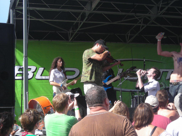 Eine Gruppe von Musikern, die verschiedene Instrumente auf einer Bühne spielen, während das Publikum vorne steht und die Performance genießt, mit einem großen schwarzen Lautsprecher links und einem grünen Banner im Hintergrund.