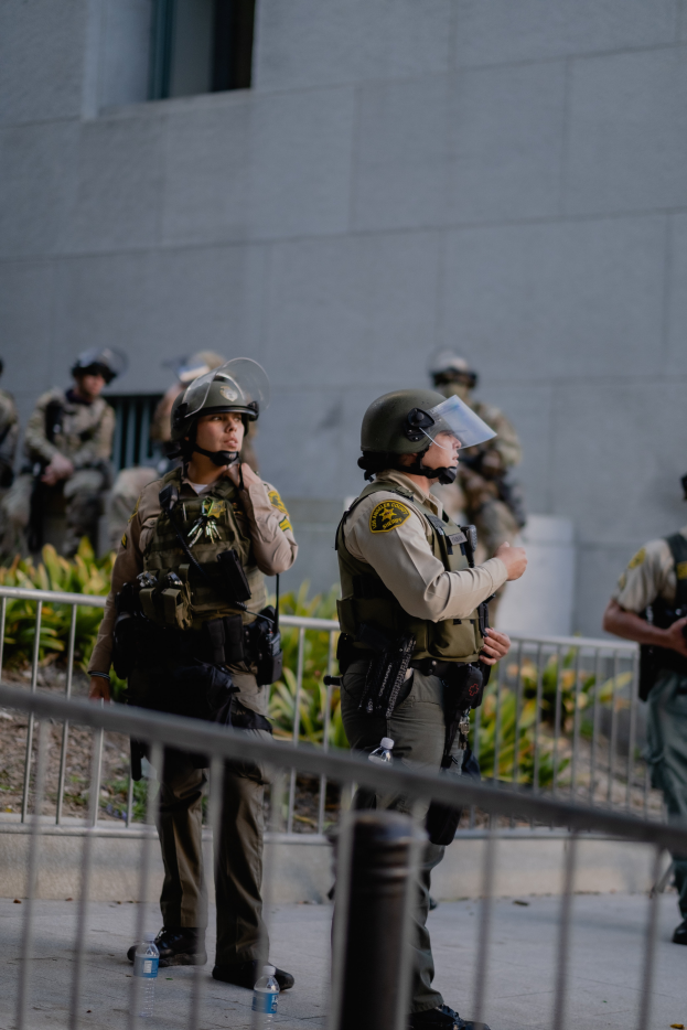Eine Gruppe uniformierter Polizisten, einige bewaffnet, steht in einer Reihe vor einem Gebäude mit einem Metallzaun im Vordergrund und Pflanzen und einer Wand im Hintergrund.