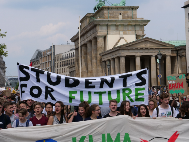 Gruppe von Schülern marschiert in Berlin mit einem bunten "Students for Future"-Schild vorbei an Gebäuden, Bäumen und Himmel.