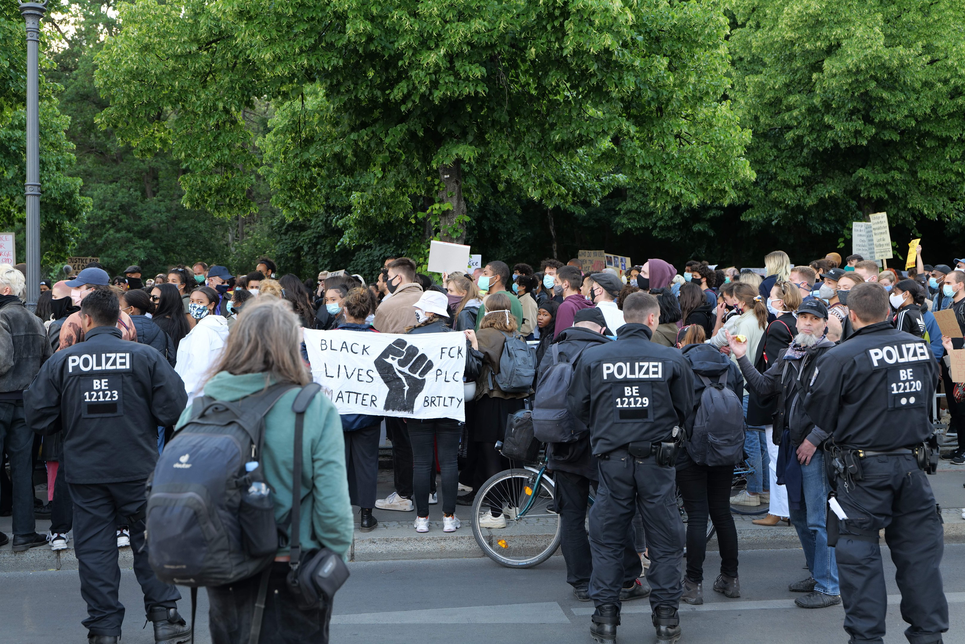 Eine große Gruppe von Menschen bei einer Black Lives Matter Demonstration in Berlin, einige mit Schildern und Mützen und Taschen, mit einem Fahrrad im Vordergrund und Bäumen und einem Pfahl im Hintergrund.