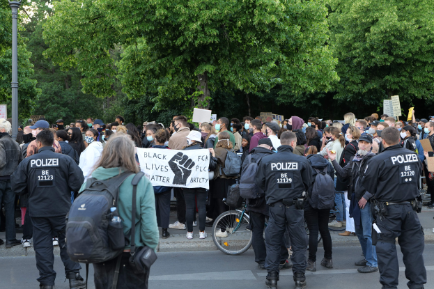 Eine große Gruppe von Menschen bei einer Black Lives Matter Demonstration in Berlin, einige mit Schildern und Mützen und Taschen, mit einem Fahrrad im Vordergrund und Bäumen und einem Pfahl im Hintergrund.