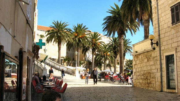 Zwei Paare spazieren auf einem Bürgersteig, der von offenen Restaurants mit Tischen und Stühlen gesäumt ist, Kokospalmen im Hintergrund und eine Backsteinwand mit Fenstergitter und Tür in der rechten Ecke.