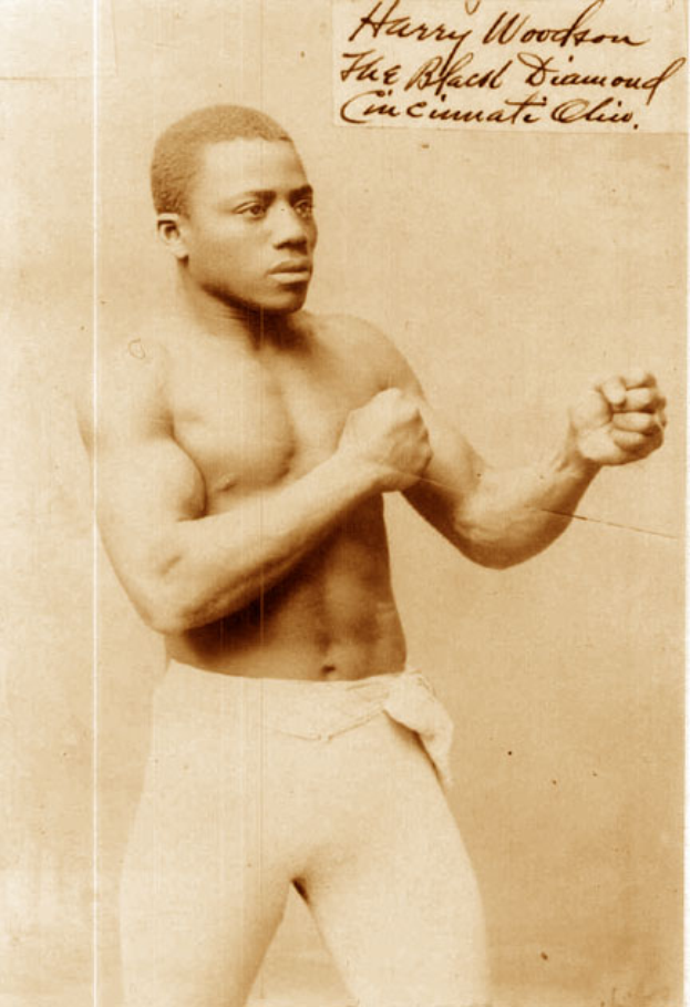 Ein altes Foto eines Mannes in einem Boxring, der Handschuhe und Shorts trägt und einen entschlossenen Gesichtsausdruck hat, kurze dunkle Haare und konzentrierte Augen, aufrecht und stolz dasteht.