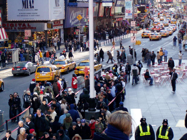 Fahrzeuge passieren eine Straße mit Fußgängern auf den Gehwegen, umgeben von Geschäften, Tischen, Stühlen, Laternen, Schildern und Ampeln.