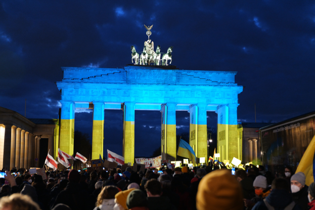 Eine Menschenmenge steht vor dem Brandenburger Tor in Berlin, Deutschland, mit Fahnen und Schildern, mit einer Fahne auf der rechten Seite und dem Torbogen mit Statuen und Säulen unter einem bewölkten Himmel.