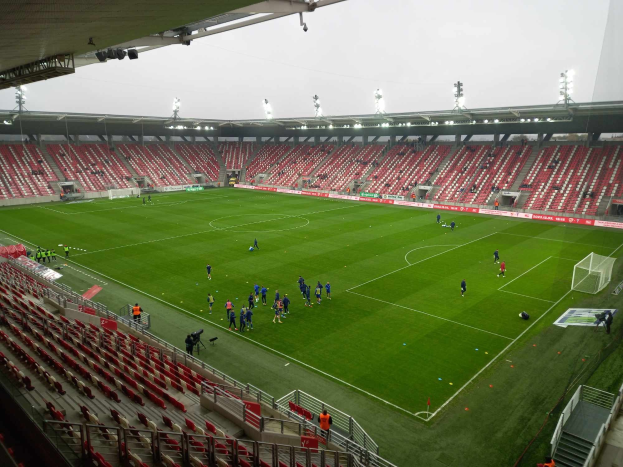 Ein Fußballfeld in einem Stadion mit Menschen, Sitzplätzen, Geländern und Beleuchtung, mit dem Himmel im Hintergrund.