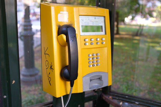 Ein gelber Münzfernsprecher auf einem Metallpfahl in der Vorderansicht, mit einer unscharfen Sicht auf einen Pfahl, Bäume und Gras durch ein Glasfenster im Hintergrund.