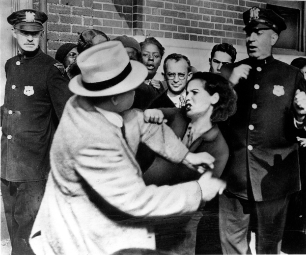 Zwei Menschen kämpfen in einem Schwarz-Wei√ü-Bild, umgeben von mehreren anderen, einige tragen Mützen, und eine Wand im Hintergrund.