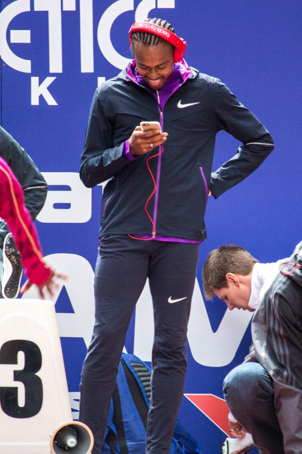 Eine Person, die mit einem Mobiltelefon in der Hand steht und lächelt, daneben eine weitere Person in Hockstellung, ein Rucksack, ein Megafon unten, eine Wand im Hintergrund und eine weitere Person, die links steht.