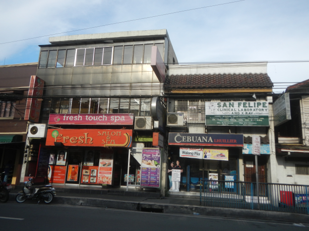 Eine belebte Stadtstraße mit verschiedenen Gebäuden, Geschäften, Fahrzeugen und elektrischer Infrastruktur, mit einem zentralen Geschäft inmitten der städtischen Aktivität.