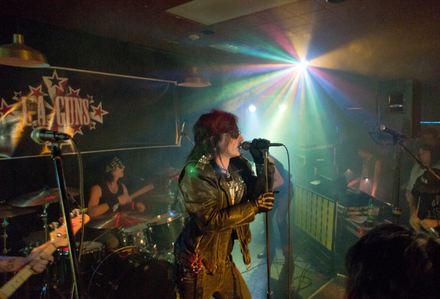 Eine Frau in einer schwarzen Jacke singt in ein Mikrofon auf der Bühne, hinter ihr spielt ein Mann mit Sonnenbrille und Stirnbinde ein Instrument; im Hintergrund sind ein "LA Guns"-Plakat und Diskoleuchten zu sehen.