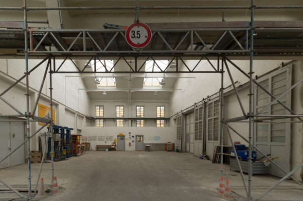 Ein großes Lager mit ausgedehnten Gerüsten, Verkehrskegeln auf dem Boden, Metallstangen, ein Schild, verschiedene Maschinen, eine Tür, Fenster, eine Wand und ein Dach mit Deckenlampen.