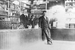 Drei Männer stehen an einem Tresen in einem Laden mit verschiedenen Gegenständen, ein buchgefülltes Regal im Hintergrund, während eines Überfalls.