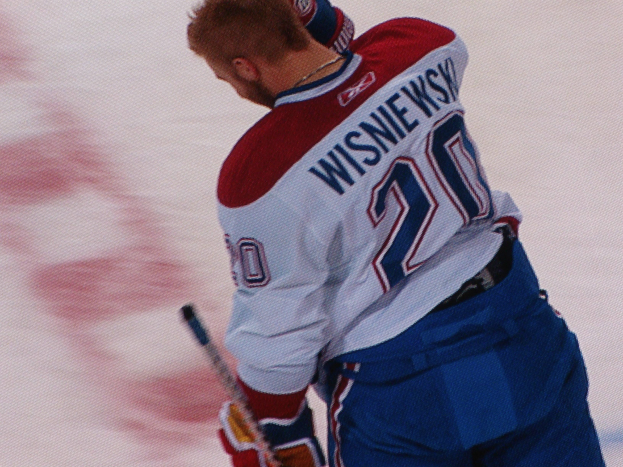 Ein Hockey-Spieler im "Wisniewski"-Trikot steht auf dem Eis, hält einen Hockey-Schläger und hat einen konzentrierten und entschlossenen Gesichtsausdruck.