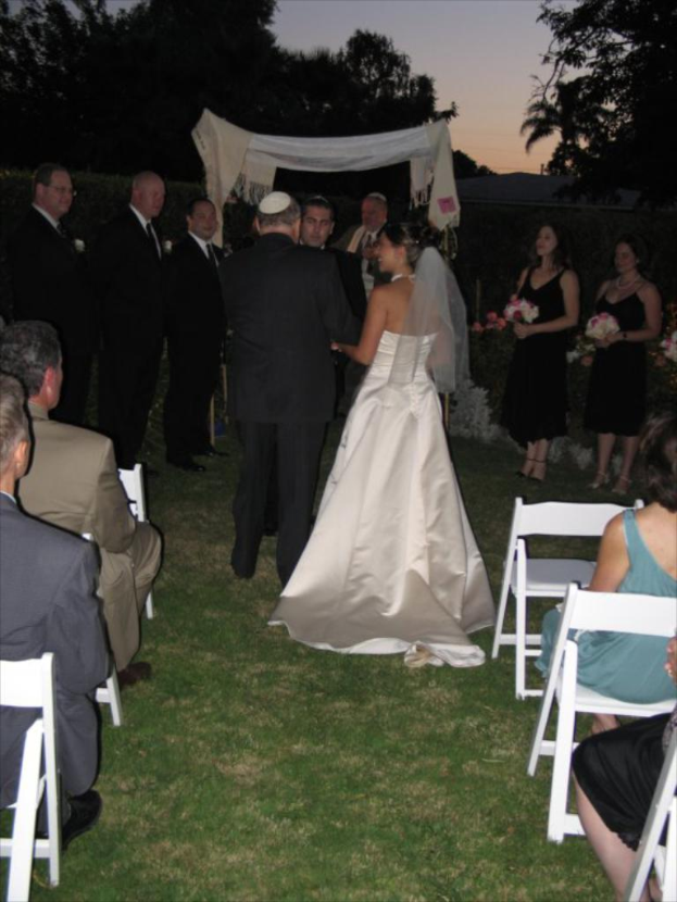 Eine Gruppe von Menschen, die bei einer Hochzeit stehen, wobei Frauen auf der rechten Seite Blumensträuße halten, während andere auf Stühlen sitzen und Bäume im Hintergrund zu sehen sind.