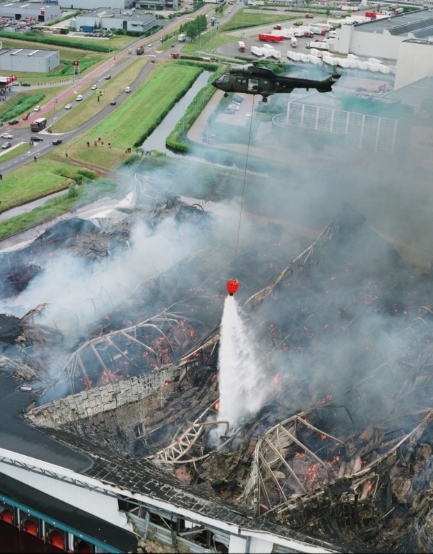 Ein Hubschrauber fliegt über ein feuerbeschädigtes Gebäude, das Rauch ausstößt, mit Fahrzeugen, Bäumen und anderen Gebäuden im Hintergrund und Text am unteren Rand.