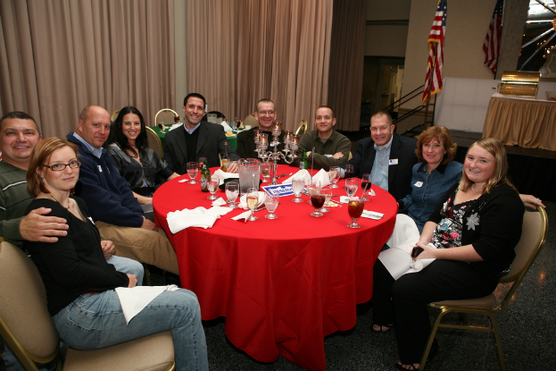 Eine Gruppe von lachenden Menschen, die an einem Tisch mit Gläsern, Papieren, Weinflaschen und Flaschen sitzen, ein Flagge auf der rechten Seite und Vorhänge an der Wand.