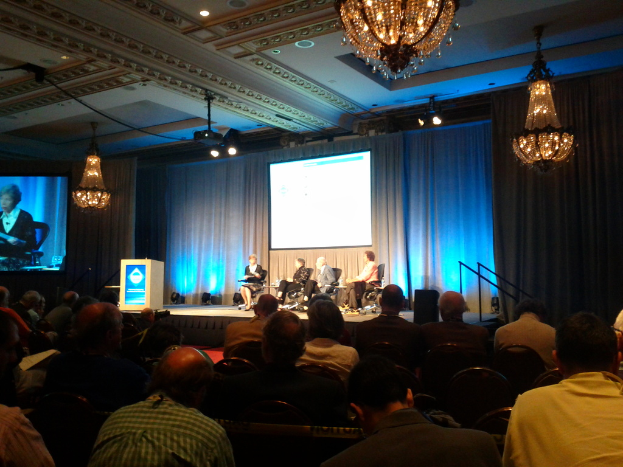 Eine Gruppe von Menschen, die auf Stühlen in einem Raum mit einer B"{u}hne, einem Podium, Scheinwerfern, Bildschirmen, Vorh"{a}ngen, Kronleuchtern, einem Redner und einem Dach sitzen.