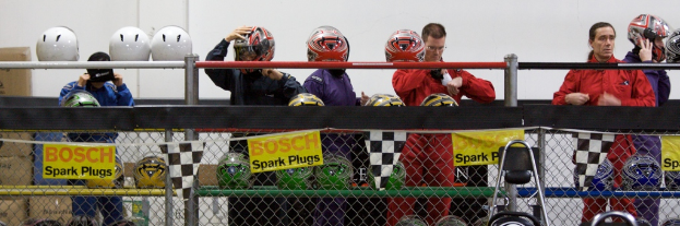 Ein Maschendrahtzaun mit Plakaten und Fahnen daran, mit ein paar Menschen im Hintergrund, einige tragen Helme, und zusätzliche Helme auf einem Ständer, vor einer Wand.