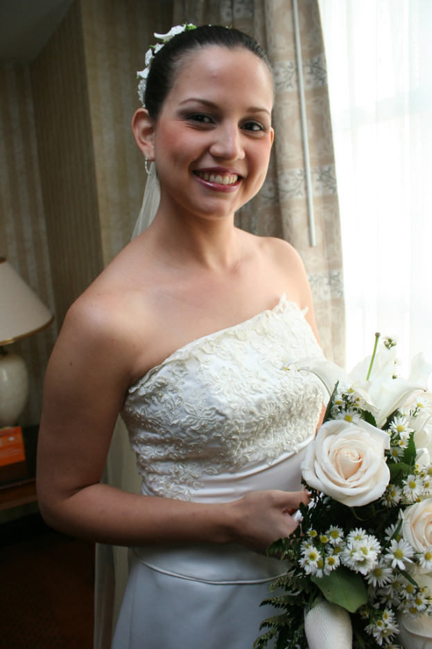 Eine Braut steht lächelnd, hält einen Strauß in der Hand, mit einem Vorhang und einer Wand im Hintergrund und einer Lampe auf einem Tisch links.