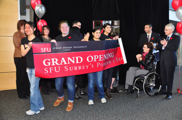 Eine Gruppe von Frauen hält ein Banner, das eine Veranstaltung ankündigt, mit zwei klatschenden Männern daneben und einer Frau im Rollstuhl, die aufgeregt aussieht, vor einem schwarzen Vorhang und Ballons.