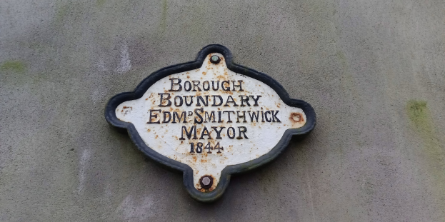 Metallplatte an der Seite eines Gebäudes mit schwarzem Hintergrund und weißer fettgedruckter Schrift, die "Borough Boundary Edm Smithwick Mayor" lautet.
