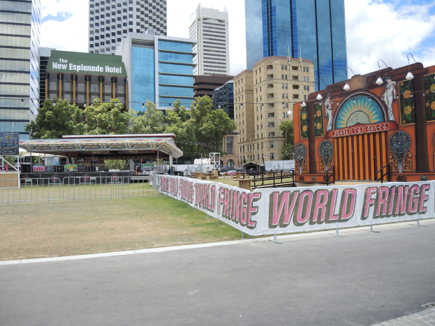 Ein Karnevalsumzug in einer Stadtstraße mit hohen Gebäuden, Bäumen und einem bewölkten Himmel im Hintergrund, eine Straße mit einem Geländer und einem Banner mit Text am unteren Rand.
