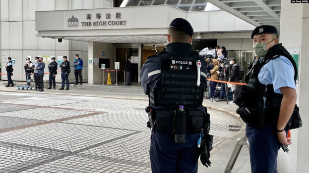 Zwei Polizisten in Uniform, Mützen und Masken stehen vor einem Gebäude am Hong Kong International Airport, umgeben von einer Gruppe von Menschen, einige halten Kameras, mit Pfählen im Hintergrund, die mit Bändern geschmückt sind.