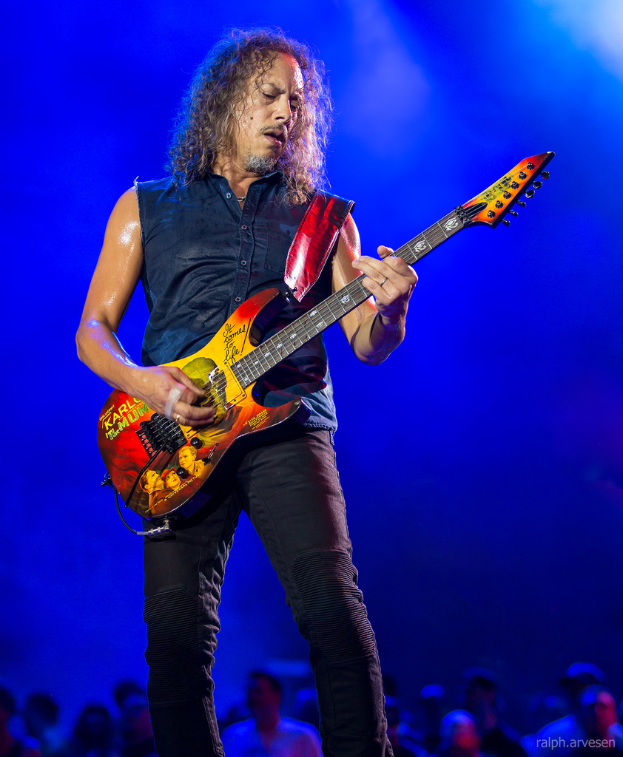 Eine Person in einem schwarzen Kleid steht in der Mitte eines musikalischen Konzerts und spielt eine Gitarre, während viele Menschen von unten zusehen.