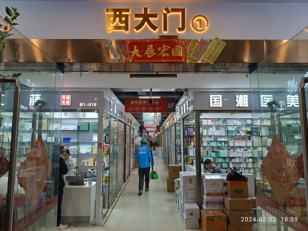 Innenseite einer chinesischen Apotheke mit Glaswänden, einer Pflanze links, Regalen voller Gegenstände, verstreuten Kartons und einem Namensschild oben.