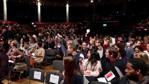 Eine Gruppe von Menschen, die auf Stühlen sitzen, Papiere halten und ID-Karten tragen, mit Säulen, Lampen, Lautsprechern und zusätzlichen Stühlen im Hintergrund.