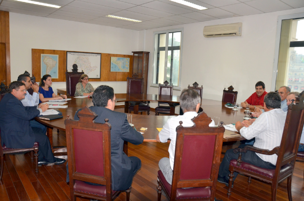 Eine Gruppe von Menschen sitzt an einem Tisch mit Papieren und Gläsern, einer Klimaanlage, einer Anschlagtafel und Karten an der Wand im Hintergrund.