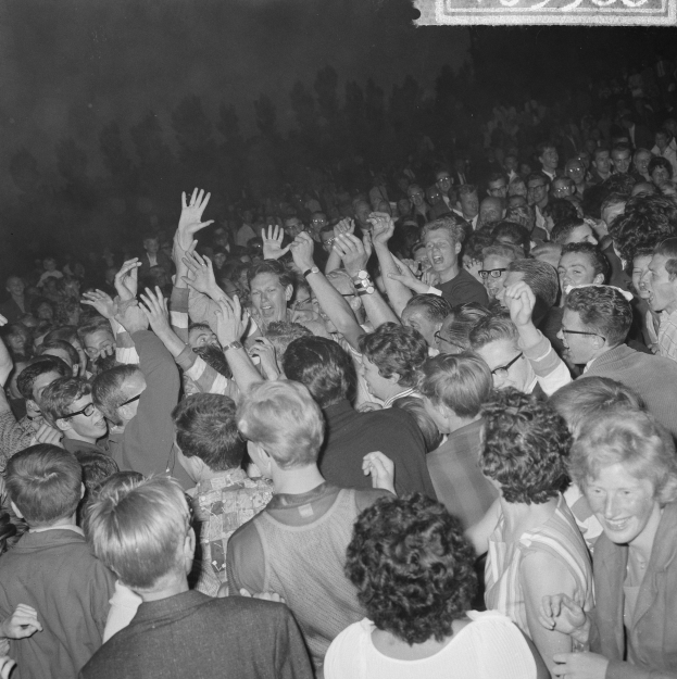 Ein Schwarz-Wei├č-Bild einer Menge auf einem Konzert mit erhobenen H├Ąnden um eine Tafel mit Text.