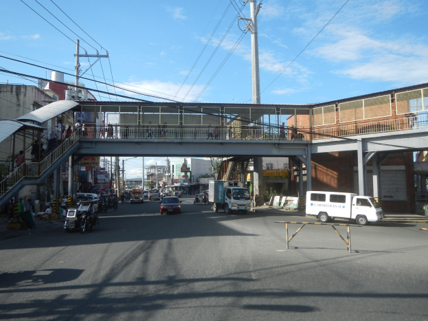 Stadtstraße mit Fahrzeugen, eine Fußgängerbrücke mit Menschen, Strommäste, Gebäude und ein bewölkter Himmel.