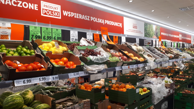 Ladeninneres mit verschiedenen Obst- und Gemüsesorten in Kartonschachteln mit Preisschildern, Wandtafeln mit Text und Bildern sowie Deckenleuchten.