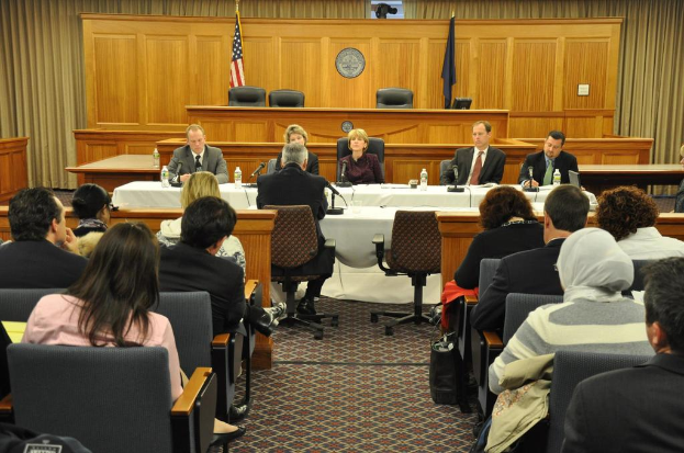 Ein Versammlungsraum mit einer Gruppe von Menschen, zwei ausländischen Flaggen, einem orangen Schreibtisch mit Wasserflaschen und einem Mikrofon.