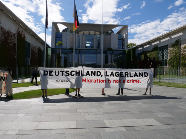 Gruppe von Menschen mit einem Banner "Deutschland, Lagerland Migration ist kein Verbrechen" vor einem Gebäude mit Fenstern, umgeben von Gras, einem Metallzaun, Pflanzen, Bäumen und Fahnenstangen unter einem bewölkten Himmel.