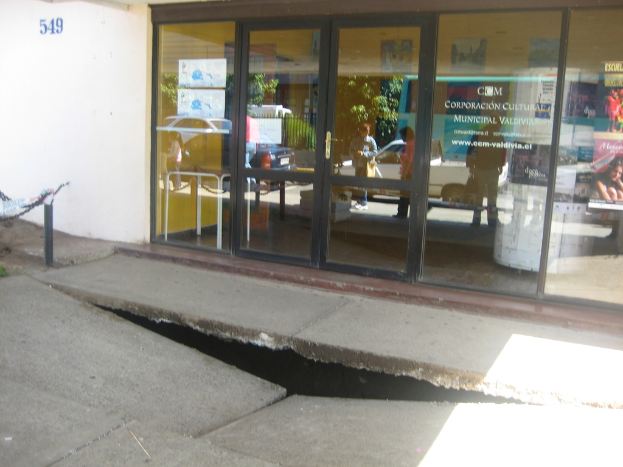 Eine gebrochene Oberfläche im Vordergrund mit einer Glastür, Wand und einem Pfahl im Hintergrund, die sich bewegende Fahrzeuge und einige stehende Personen spiegeln.
