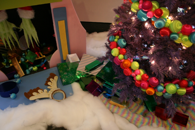 Innen: Ein geschmückter Weihnachtsbaum mit Lichterkette und Schmuck steht rechts, mit Geschenkboxen und Dekorationen in der Mitte, einer Hütte mit Dekorationen oben links und Baumwolle und Wolle unten.