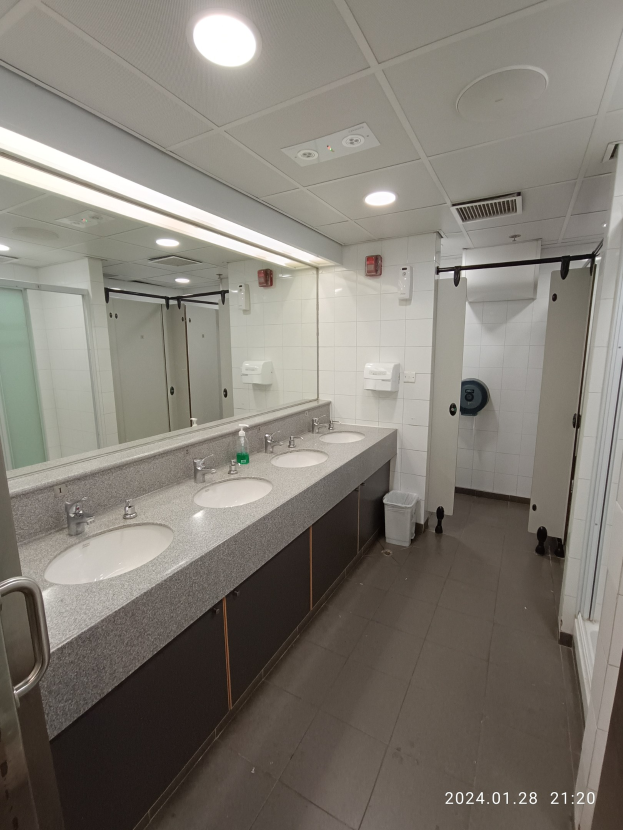 Öffentliche Toilette mit zwei Waschbecken, einem großen Spiegel, Wasserhähnen, einem Bodenmüllbehälter, einer Tür mit Griff und Deckenbeleuchtung.