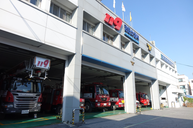 Feuerwache mit mehreren Feuerwehrautos vor dem Gebäude mit Fenstern, Säulen und Namensschildern, mit Fahnen im Vordergrund und anderen Gebäuden im Hintergrund und einem klaren blauen Himmel.