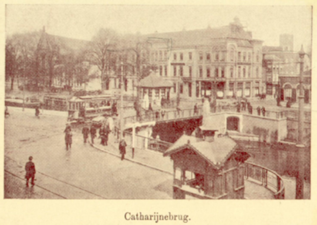 Ein altes Foto einer Stra├če mit Menschen, umgeben von Geb├Ąuden, B├Ąumen, Masten und einer ├Überquerung, mit Text am unteren Rand.