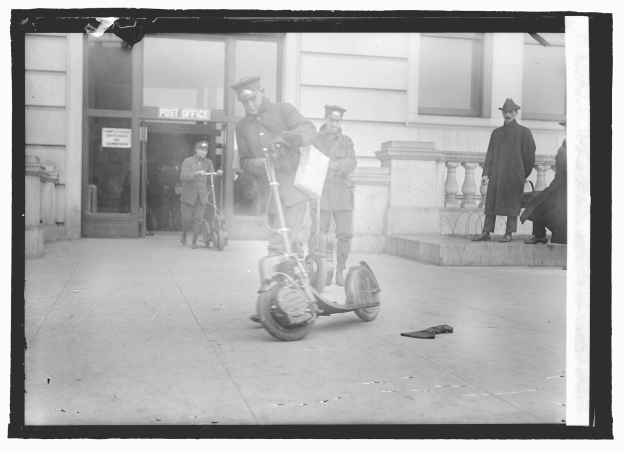 Schwarz-weißes Foto eines Mannes mit Mütze auf einem E-Scooter auf einem Gehweg, umgeben von anderen Menschen, darunter ein Radfahrer, und einem Gebäude mit einer Tür und einem Schild im Hintergrund.