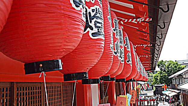 Rote chinesische Laternen h√§ngen √ľber einer Menschenmenge, mit bunten Papieren und Geb√§uden im Hintergrund, unter einem Himmel mit B√§umen.