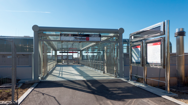 Ein Gehweg mit Geländern und Pfosten führt zu einem Bushaltestellenschild, mit Texttafeln an den Seiten und Gebäuden im Hintergrund unter einem klaren blauen Himmel.