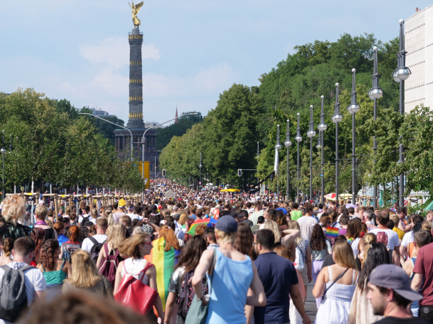 Eine große Menschenmenge mit Taschen geht eine Straße in Berlin, Deutschland, entlang, gesäumt von Laternenpfählen, Bäumen, einem Turm mit einer Statue darauf, Gebäuden und einem bewölkten Himmel im Hintergrund.