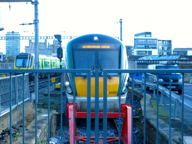 Zwei Züge auf Schienen mit einem Zaun im Vordergrund, Pfähle mit einem, der ein Signallicht hat, ein Auto auf der Straße rechts, Gebäude im Hintergrund und Himmel oben.