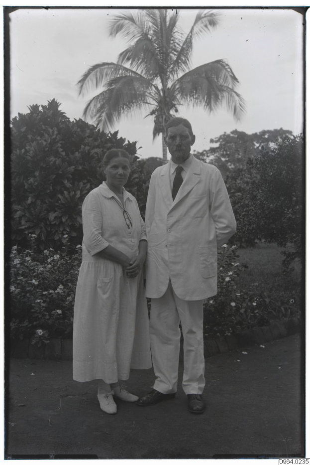 Ein altes Schwarz-Weiß-Foto eines Mannes im Anzug und einer Frau, die nebeneinander vor einer Palme stehen, mit Bäumen und einem klaren Himmel im Hintergrund.