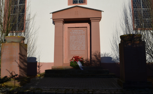 Ein Gebäude mit Fenstern, ein Denkmal mit Text, ein Blumenstrauß auf dem Boden, Topfpflanzen, eine Baumgruppe und ein bewölkter Himmel, mit einem Grab und einem Kranz davor.